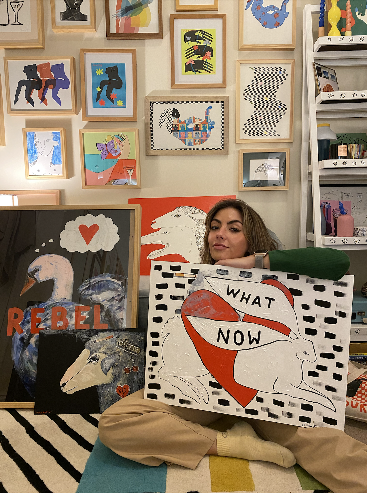 image of Elena holding the what now canvas, with other pieces of her art hanging on the wall behind her.